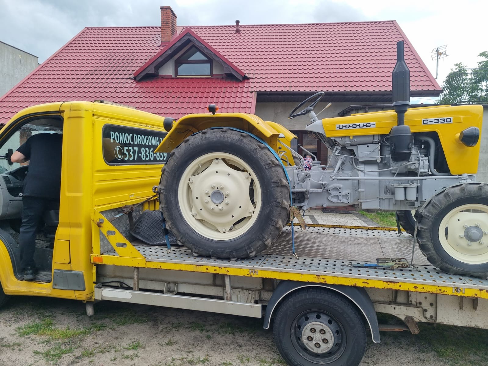 Żółta laweta z napisem 'Pomoc Drogowa' przewozi żółto-szary traktor Ursus C-330 na tle domu z czerwoną dachówką.