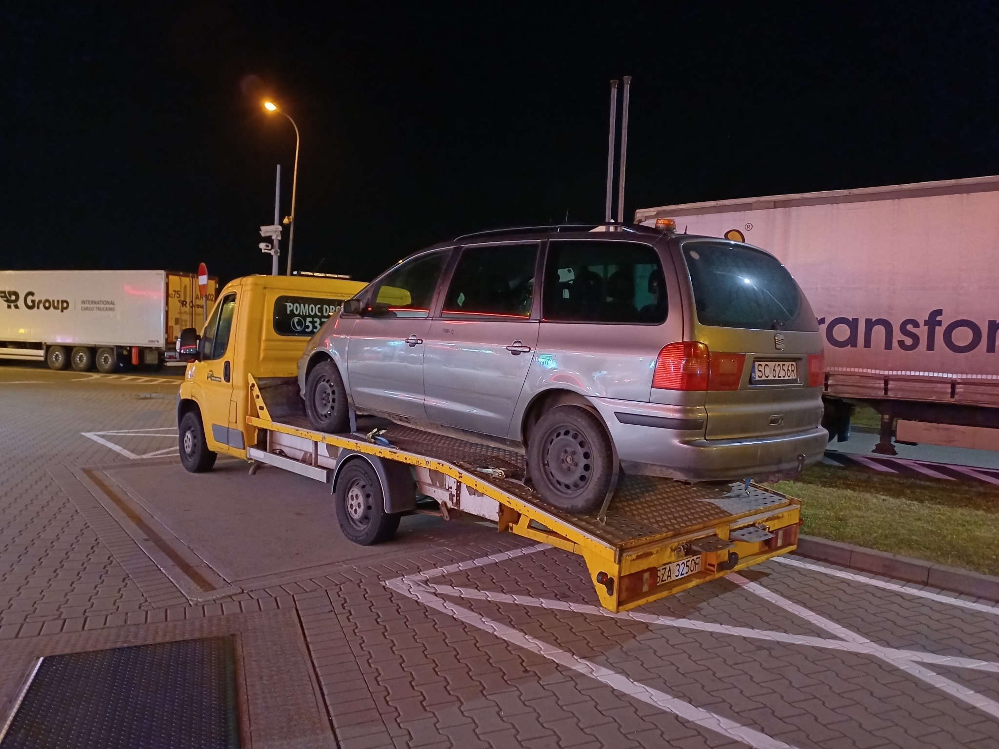 Żółta laweta z szarym samochodem osobowym na platformie, sfotografowana nocą na parkingu, w tle widoczne naczepy TIR