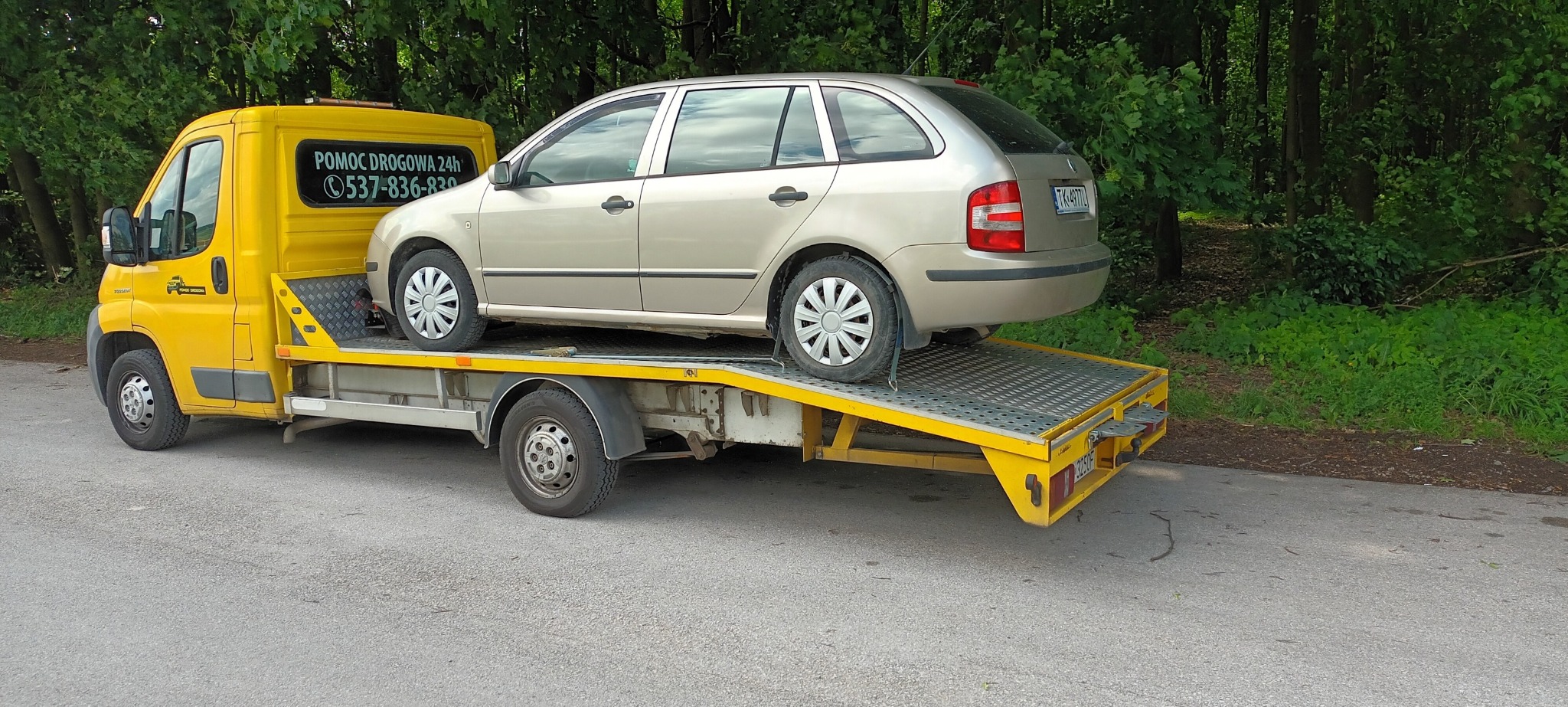 Żółta laweta z beżowym samochodem osobowym Skoda Fabia kombi na platformie, widoczne tablice rejestracyjne z Polski, tło z zielenią drzew.