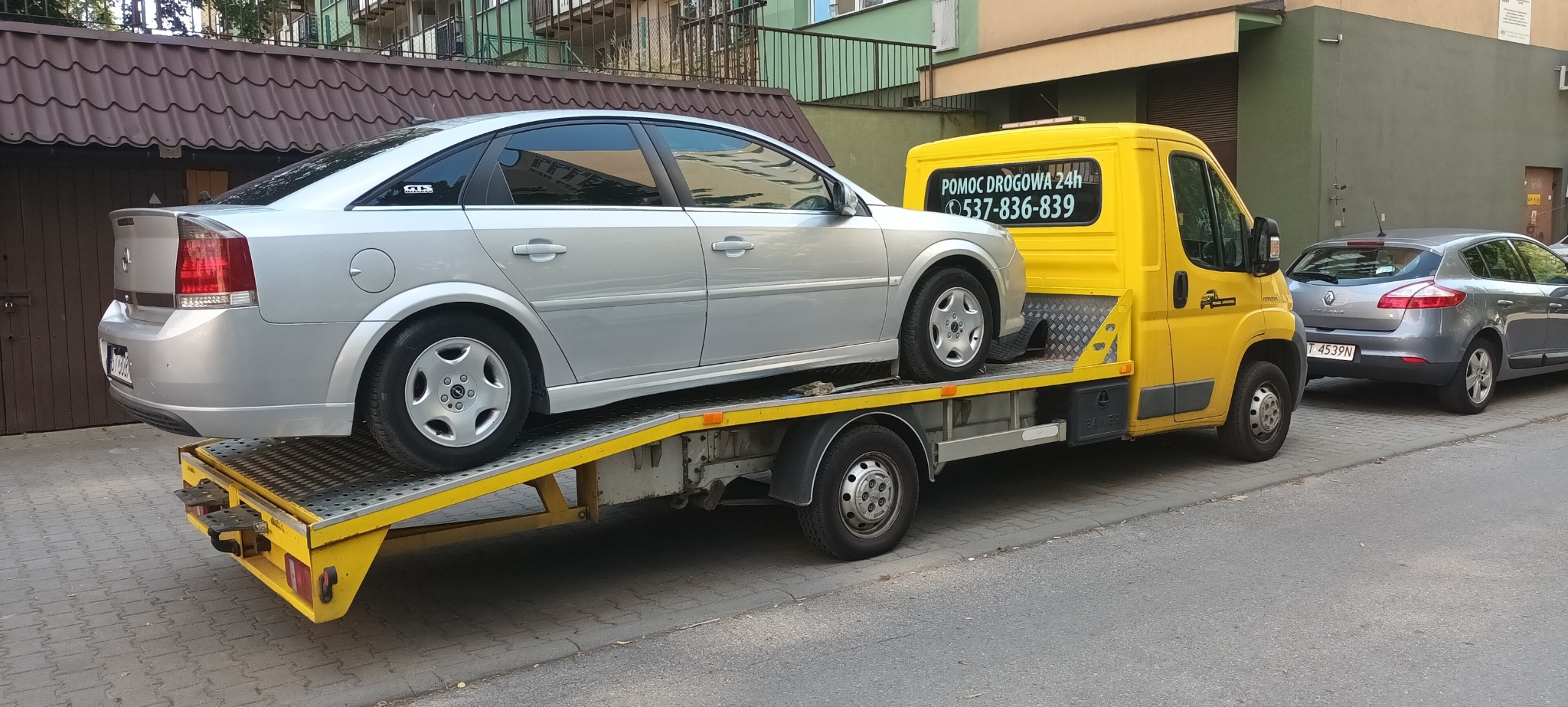 Srebrny Opel Vectra na żółtej lawecie na tle miejskiej ulicy, widoczny numer telefonu 'Pomoc Drogowa 24h'