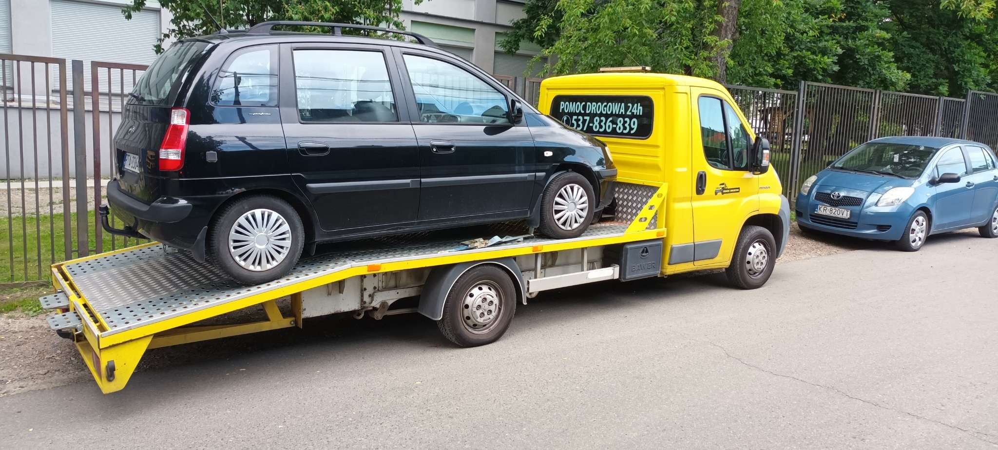 Czarny samochód osobowy załadowany na żółtą lawetę z napisem 'Pomoc Drogowa 24h' i numerem telefonu, obok zaparkowany niebieski samochód.