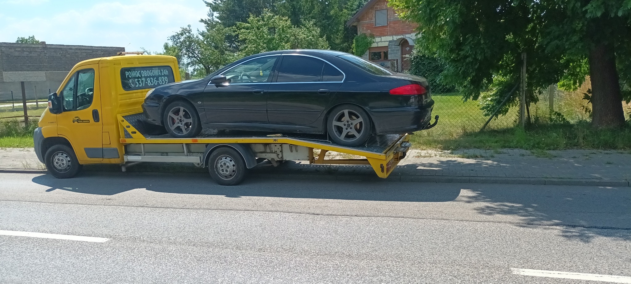 Żółta laweta z czarnym samochodem osobowym na platformie, zaparkowana na poboczu drogi, widok z boku w słoneczny dzień.