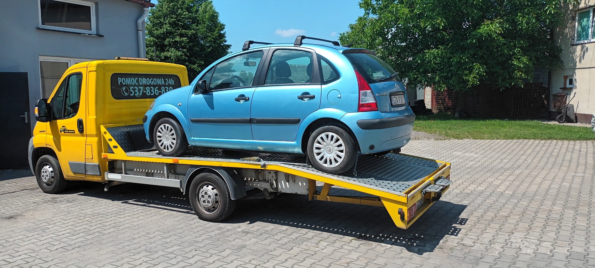 Żółta laweta z niebieskim samochodem Citroen C3 na platformie, widok z tyłu pojazdu na tle szarego budynku i zieleni.