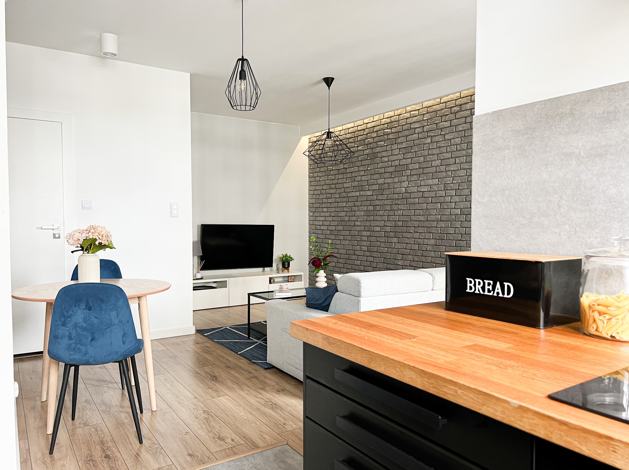 Jasne, nowocześnie urządzone wnętrze mieszkania z drewnianym stołem, niebieskimi krzesłami, szarą sofą i dekoracyjną ścianą z cegły.