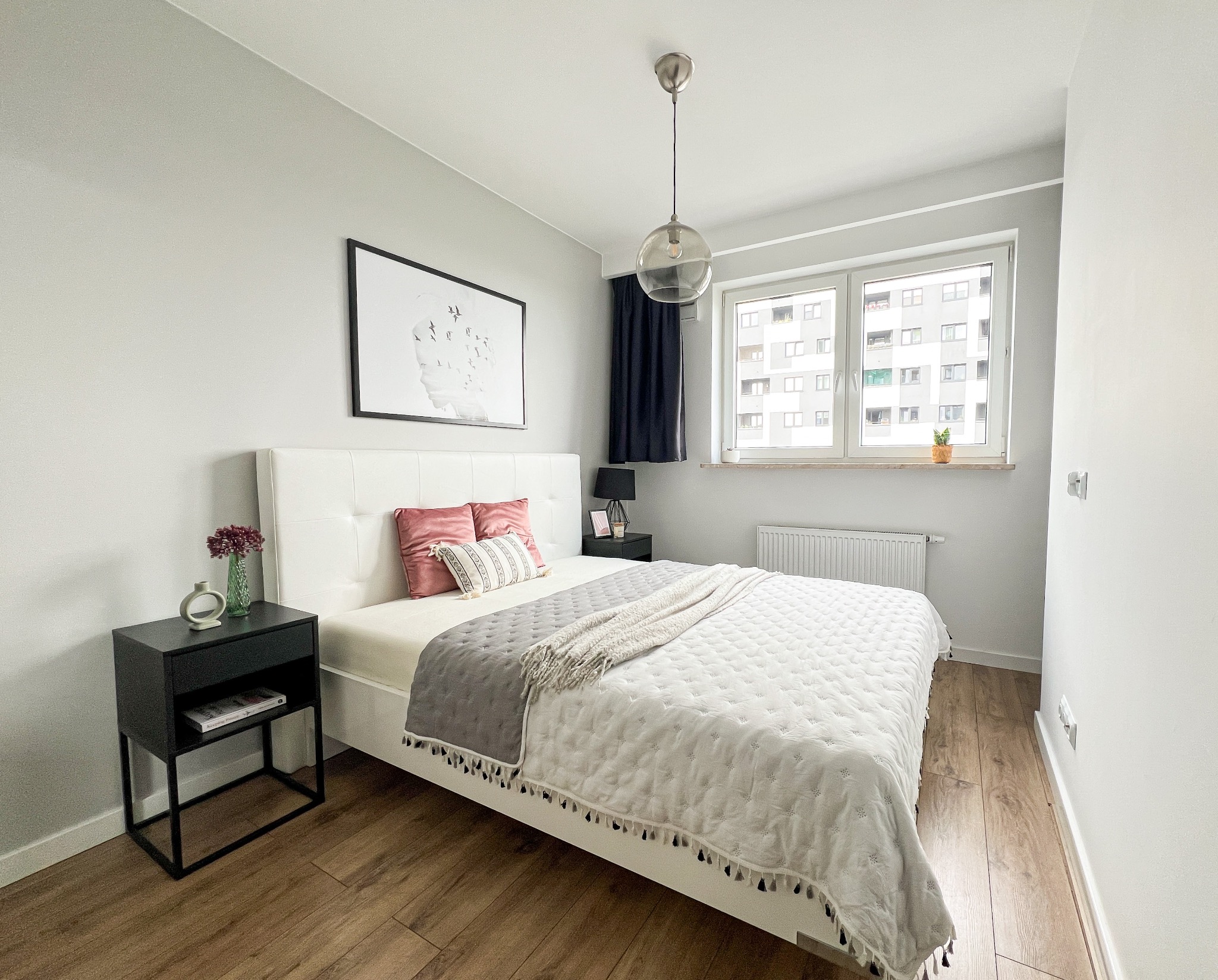 Jasna sypialnia z białym łóżkiem, szarą narzutą i drewnianą podłogą, widok na budynek mieszkalny za oknem, minimalistyczny wystrój.