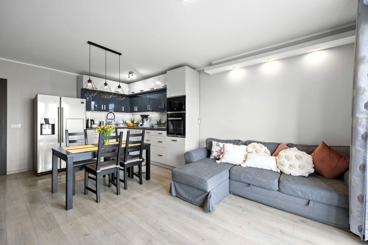 Jasne wnętrze mieszkania z szarą sofą narożną, drewnianym stołem jadalnianym z czarnymi krzesłami i nowoczesną kuchnią z białymi i granatowymi szafkami.