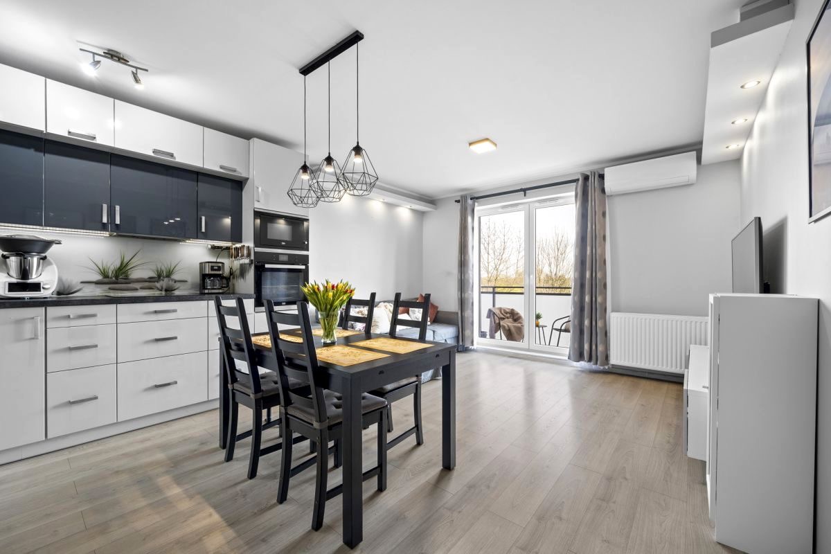 Jasne, przestronne wnętrze mieszkania z aneksem kuchennym, czarnym stołem jadalnym z bukietem tulipanów i wyjściem na balkon.