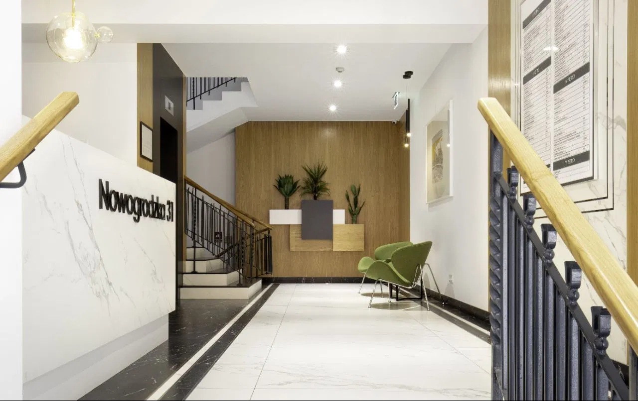Wnętrze nowoczesnego holu wejściowego z marmurową recepcją z napisem 'Nowogrodzka 31', roślinami w donicach, zielonymi fotelami i schodami z czarną balustradą.