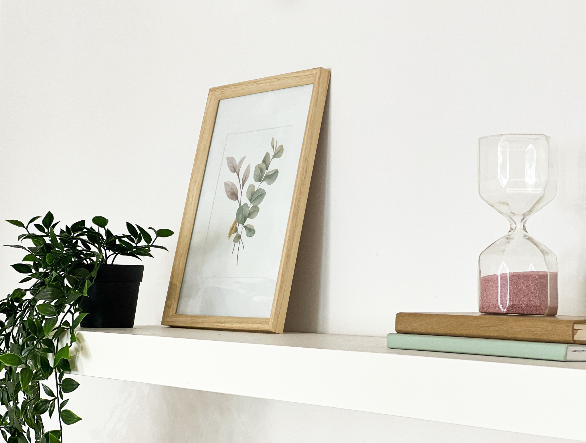 Dekoracja wnętrza: roślina w czarnej doniczce, ramka z botanicznym motywem oraz klepsydra z różowym piaskiem na stosie książek, na białej półce.