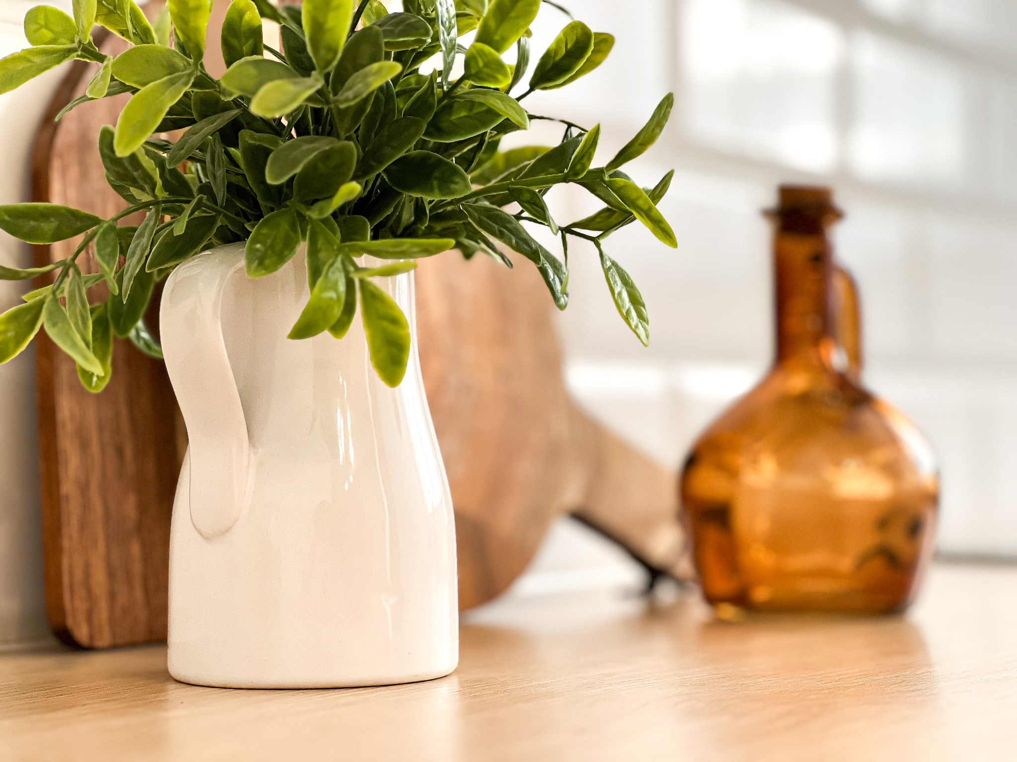 Dekoracja kuchenna: Biały dzbanek z zielonymi gałązkami na drewnianym blacie, w tle drewniana deska do krojenia i brązowa butelka.