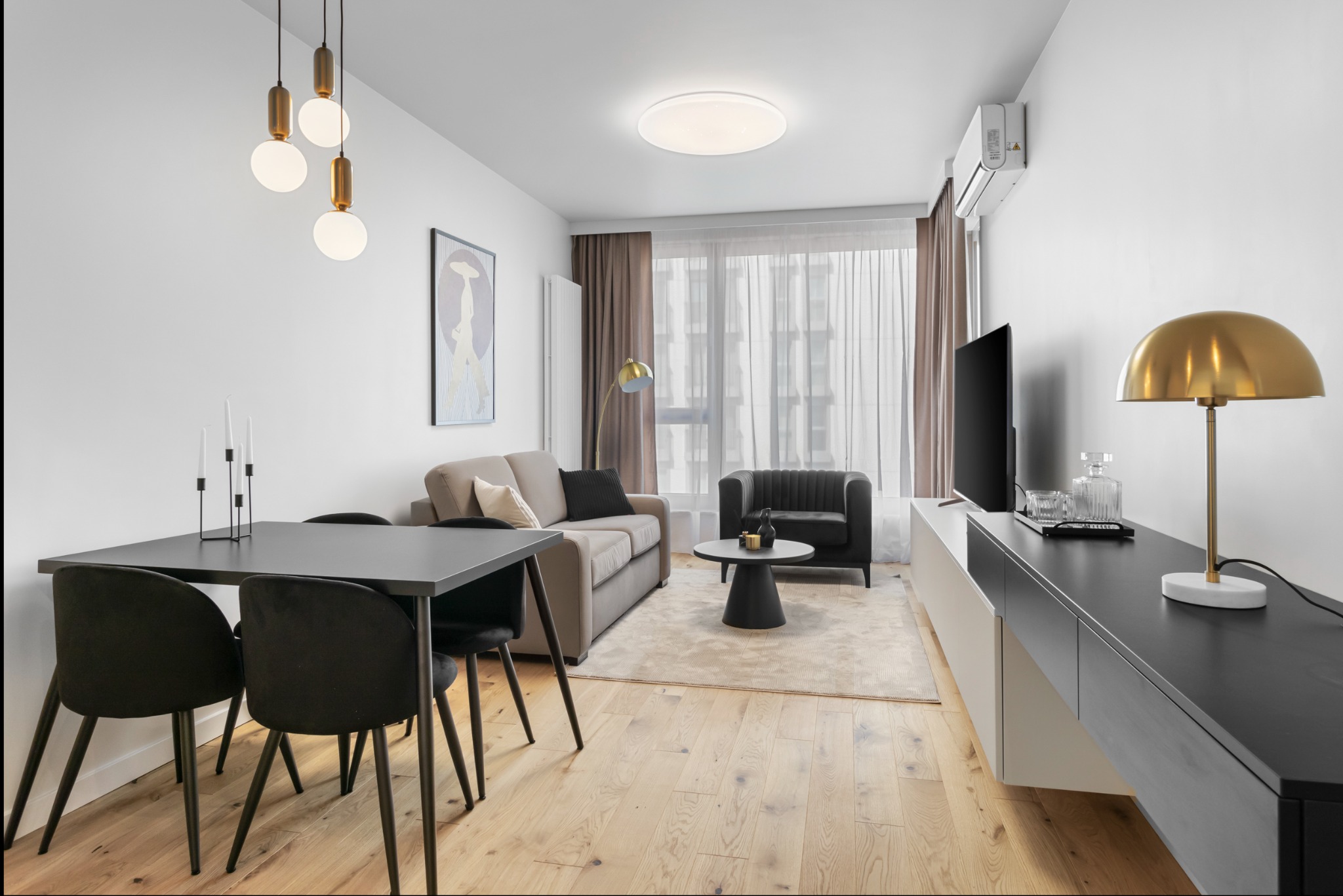 Jasne wnętrze mieszkania z drewnianą podłogą, szarym stołem i krzesłami, beżową sofą i czarnym fotelem, oświetlone wiszącymi lampami i lampą stołową z kloszem w kolorze złota.