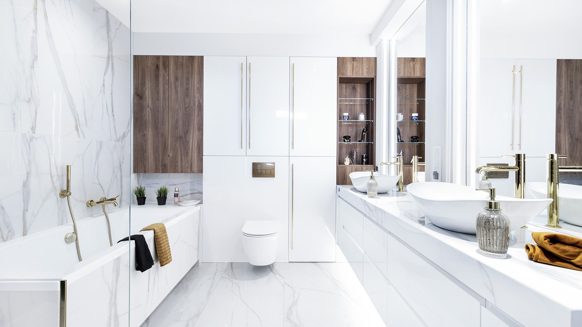 Nowoczesna, jasna łazienka z białymi marmurowymi płytkami, złotą armaturą, wiszącą toaletą i umywalkami nablatowymi. W tle szafki z białymi frontami i drewnianymi wstawkami.