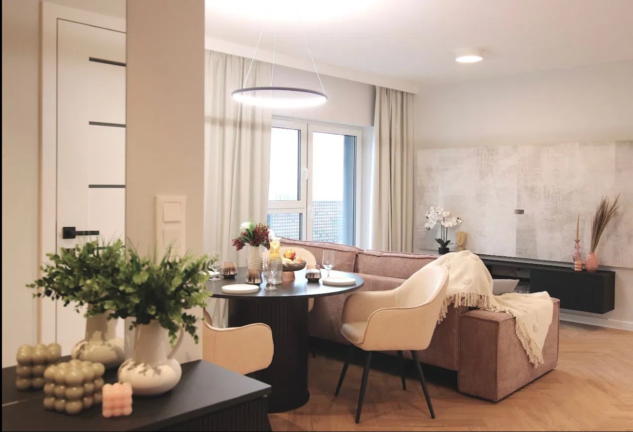 Jasne wnętrze salonu z okrągłym stołem, beżowymi krzesłami, sofą z korduroy i dekoracjami w pastelowych kolorach, oświetlone nowoczesną lampą wiszącą.
