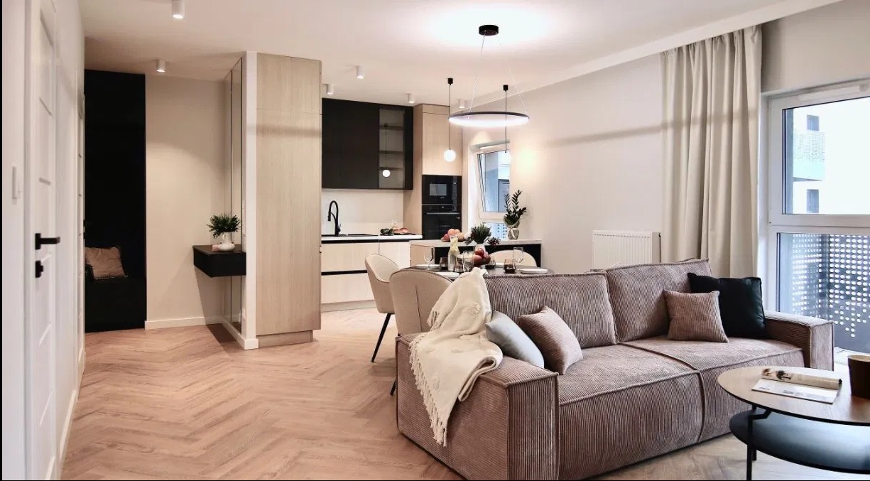 Jasne, przestronne wnętrze mieszkania z parkietem w jodełkę, obejmujące salon z sofą w odcieniach brązu, jadalnię z owocami na stole i aneks kuchenny z czarnymi akcentami.