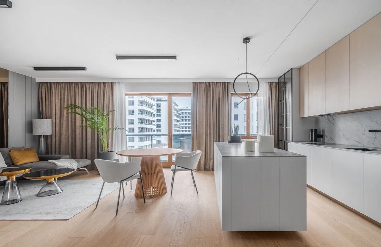 Jasne, przestronne wnętrze apartamentu z otwartą kuchnią, okrągłym stołem z ażurową podstawą i widokiem na budynki mieszkalne za oknem.