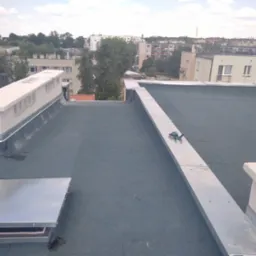 Płaski dach pokryty papą termozgrzewalną z elementami wentylacyjnymi, obróbkami blacharskimi i butelką wody, na tle zabudowy miejskiej.