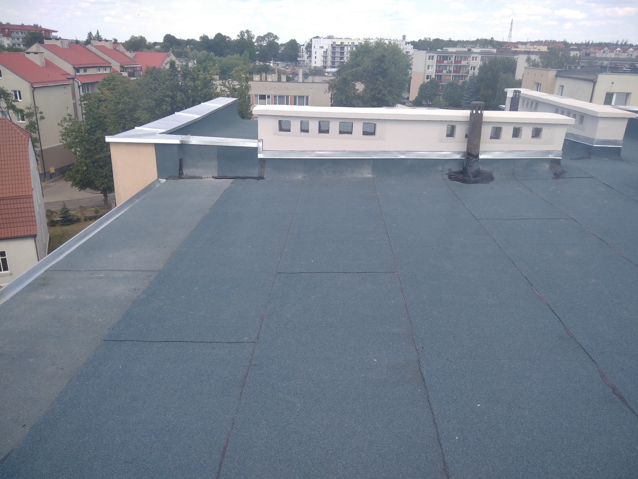 Płaski dach pokryty papą termozgrzewalną, widoczne kominki wentylacyjne i obróbki blacharskie, w tle zabudowa miejska.