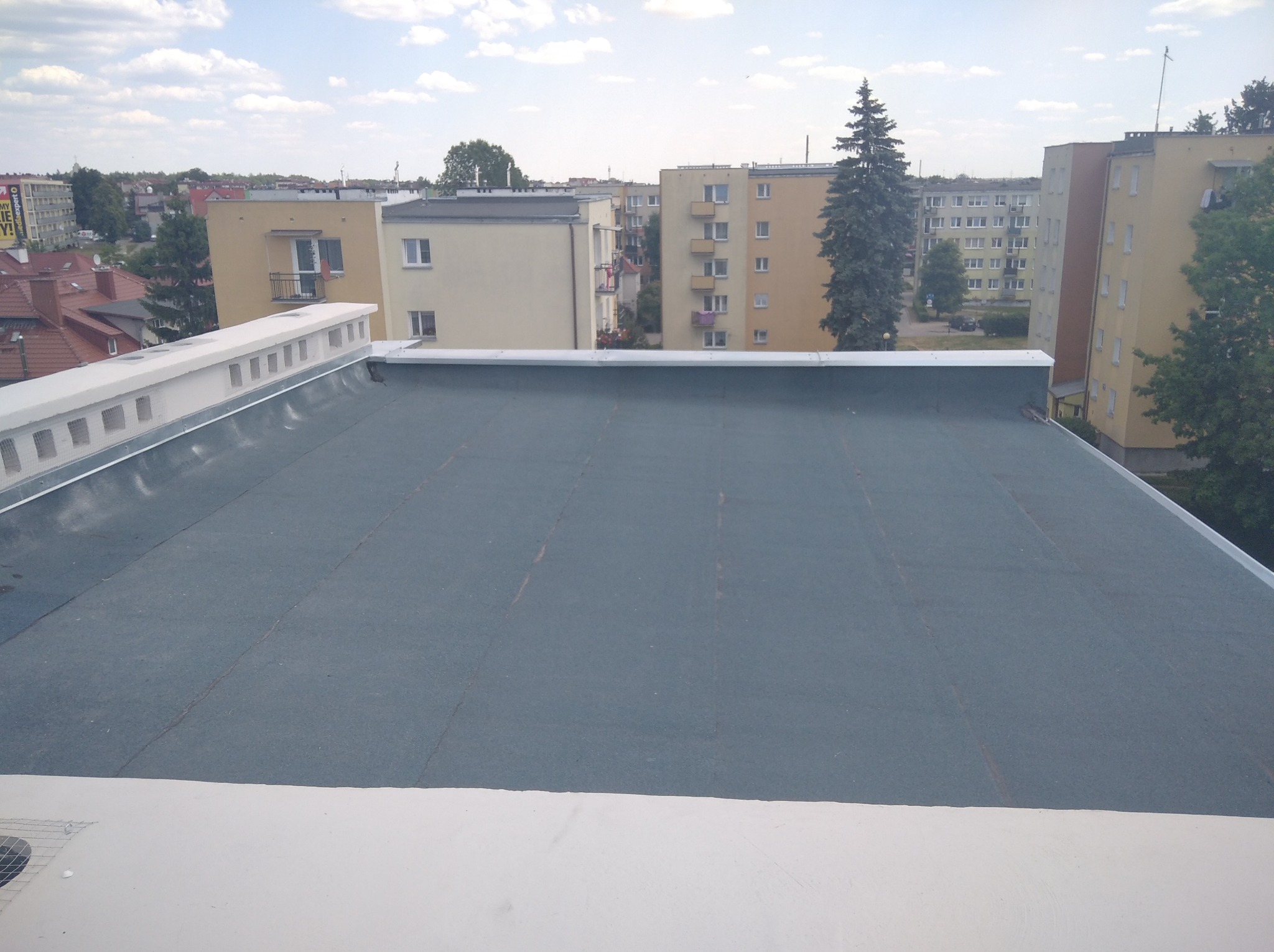 Płaski dach pokryty ciemnoszarą papą termozgrzewalną, z widocznymi zgrzewami, obrzeża wykończone białym elementem, w tle zabudowa miejska i błękitne niebo.