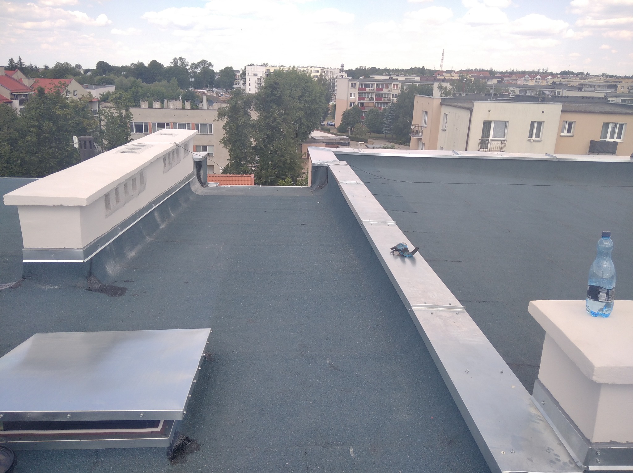 Płaski dach pokryty papą termozgrzewalną z elementami wentylacyjnymi, obróbkami blacharskimi i butelką wody, na tle zabudowy miejskiej.