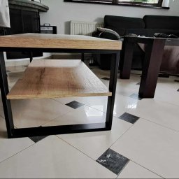Zbyszek Molenda - Stolik kawowy z drewnianym blatem i metalową ramą, na tle salonu z sofą i stolikiem. Widoczne nogi osoby w tle. Nowoczesny design, jasne wnętrze.