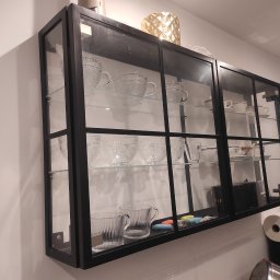 Zbyszek Molenda - Czarna, metalowa witryna z przeszklonymi drzwiami, zamontowana na ścianie. Wewnątrz szklane półki z filiżankami i dekoracjami. Minimalistyczny design, widoczne mocowania.