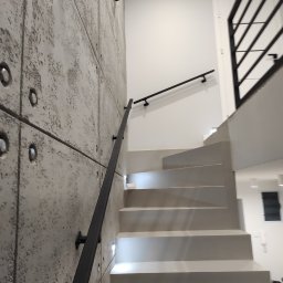 Zbyszek Molenda - Nowoczesne schody z szarą betonową ścianą i czarną, minimalistyczną poręczą. Widoczne oświetlenie schodów i fragment balustrady na górze.