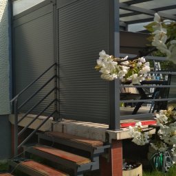 Zbyszek Molenda - Szara zabudowa balkonu z roletami i metalową konstrukcją, widoczna z zewnątrz, z drewnianymi schodami i kwitnącą gałęzią drzewa w pierwszym planie.