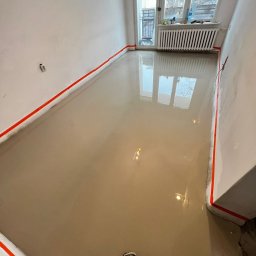 ALEX MALARZ Oleksii Tsiluiko - Wylana, mokra posadzka samopoziomująca w pokoju z oknem balkonowym. Ściany zabezpieczone taśmą. Widoczne odbicie okna w mokrej powierzchni.