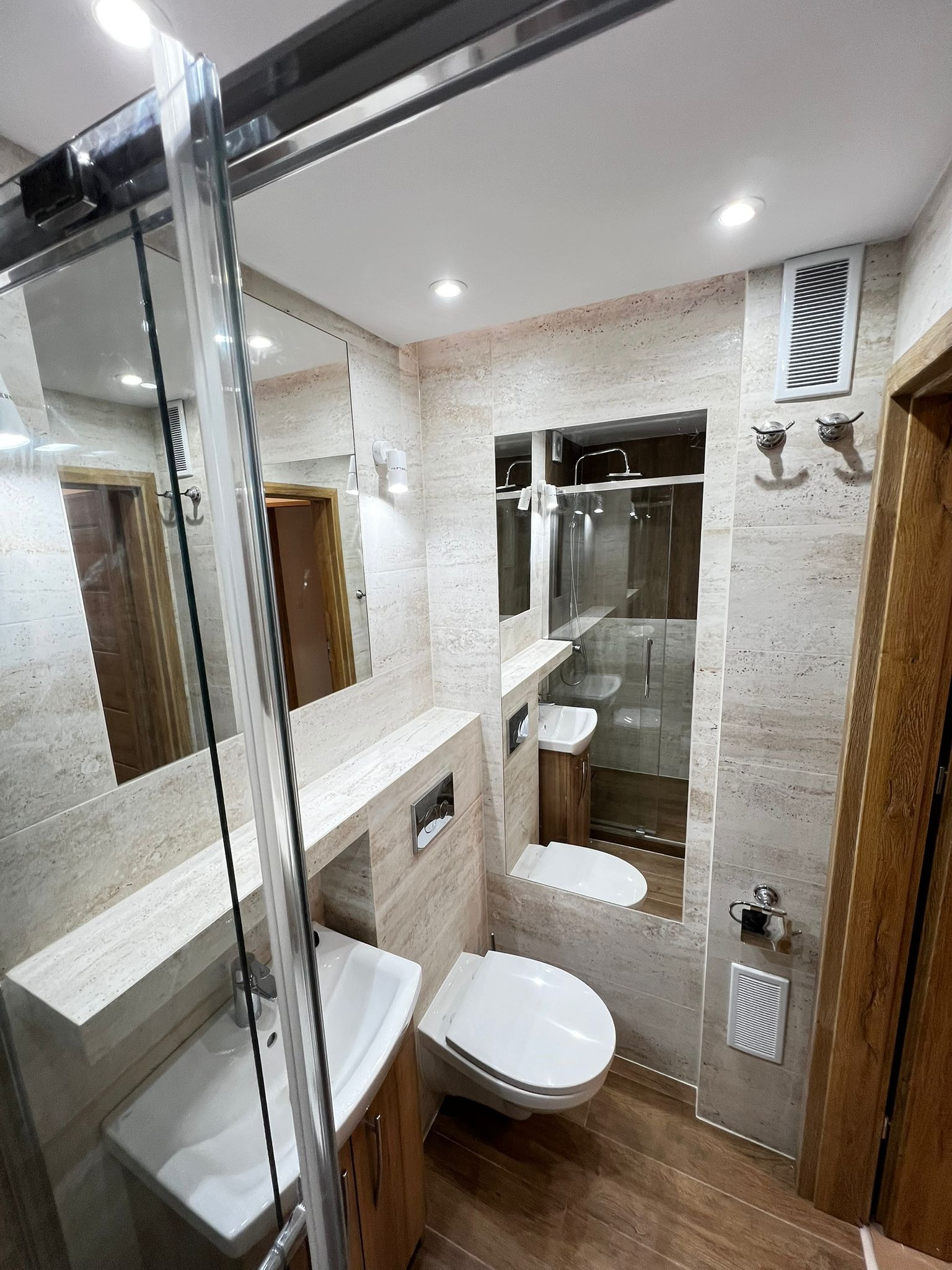 Wykończona łazienka w beżowych tonacjach z kabiną prysznicową, umywalką w zabudowie i lustrami. Widoczne drewniane akcenty i oświetlenie punktowe.