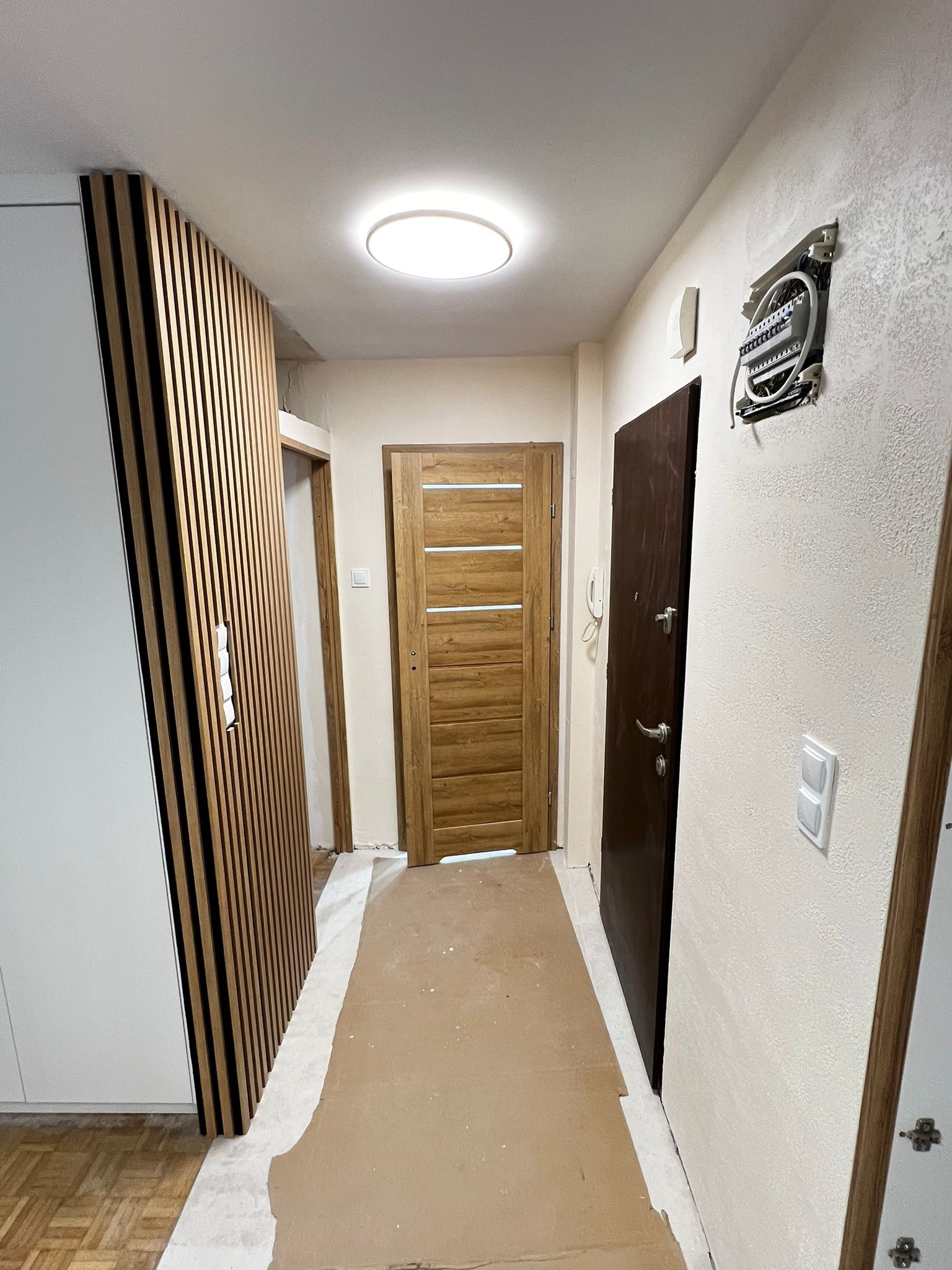 Korytarz po remoncie: drewniane lamele na ścianie, nowe drzwi wewnętrzne i wejściowe, zabezpieczona podłoga. Widoczna skrzynka elektryczna przed montażem.