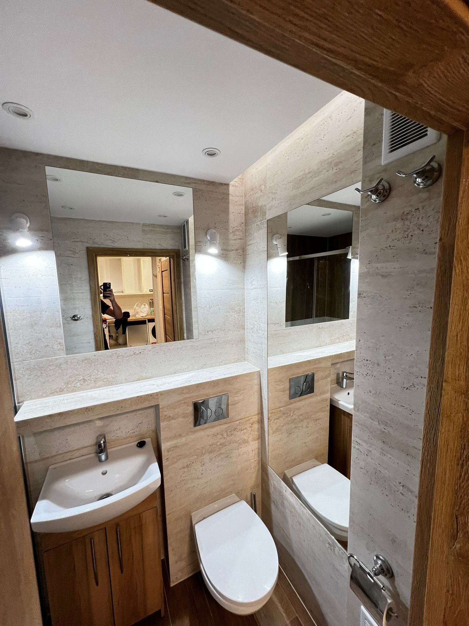 Wykończona łazienka w beżowych płytkach imitujących kamień, z drewnianą szafką pod umywalką i dużym lustrem. Widoczne detale: chromowane wieszaki i przyciski spłuczki.