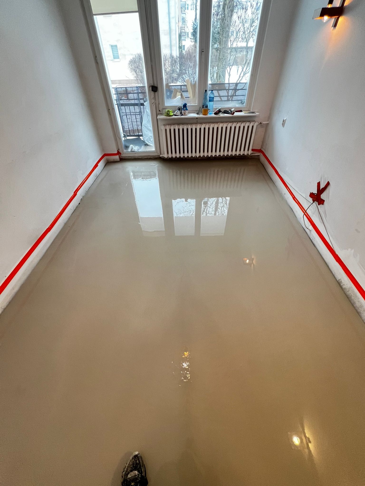 Wylana, świeża wylewka samopoziomująca w pokoju z oknem balkonowym. Czerwona taśma oddziela ściany. Widoczne odbicie okna w mokrej powierzchni.