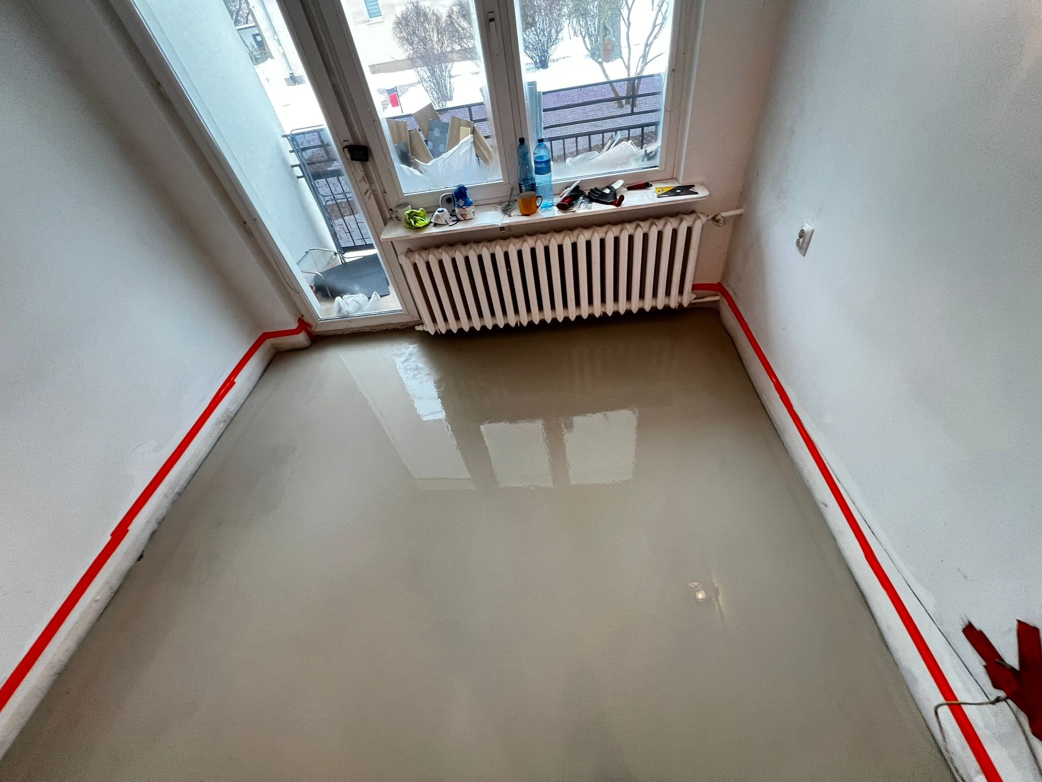 Wylany samopoziomujący podkład podłogowy w pokoju z grzejnikiem i oknem balkonowym. Czerwona taśma oddziela ściany. Widoczne narzędzia i butelka wody na parapecie.