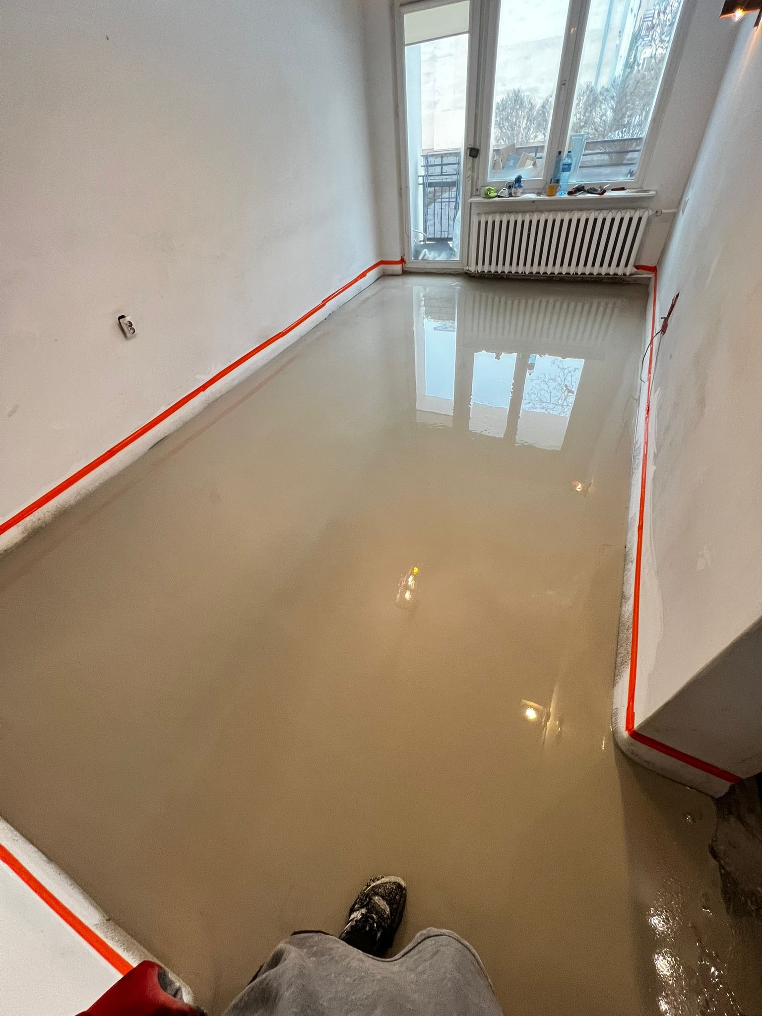 Wylana, mokra posadzka samopoziomująca w pokoju z oknem balkonowym. Ściany zabezpieczone taśmą. Widoczne odbicie okna w mokrej powierzchni.