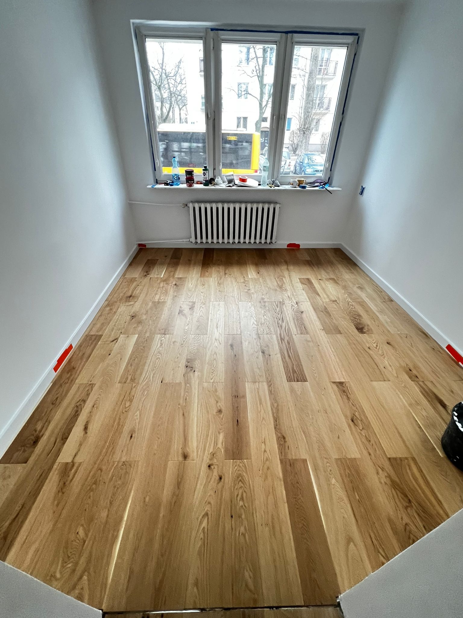 Jasna, drewniana podłoga ułożona w pokoju z białymi ścianami. Widoczne zabezpieczenia taśmą malarską przy listwach. Za oknem widok na budynki i przejeżdżający autobus.