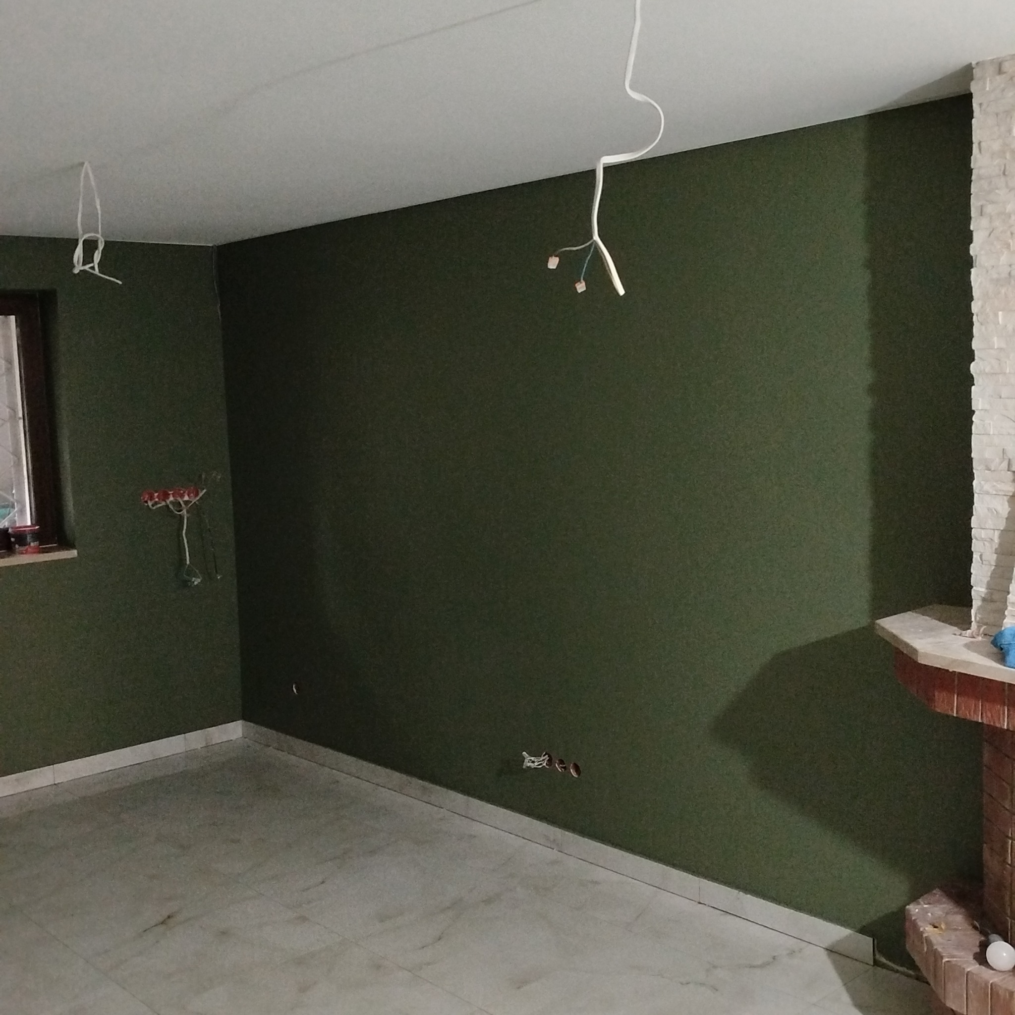 Pokój po malowaniu na oliwkowy kolor z widocznymi przewodami elektrycznymi i fragmentem kominka z cegły. Podłoga wyłożona płytkami w jasnym odcieniu.