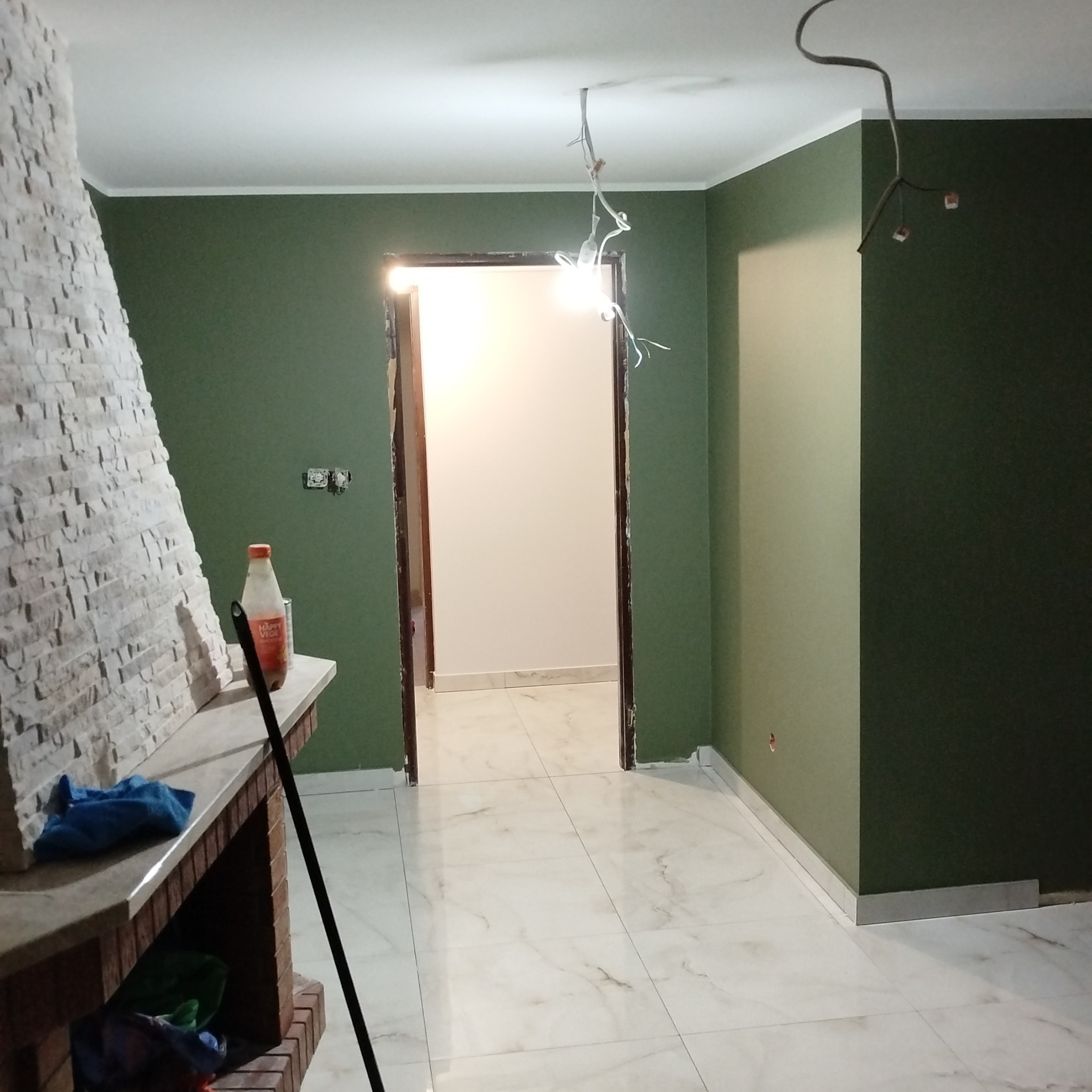 Wnętrze po malowaniu ścian na oliwkowy kolor, widoczny kominek z kamienia, biała podłoga z połyskiem i wystające kable oświetleniowe. Widok na sąsiednie pomieszczenie.