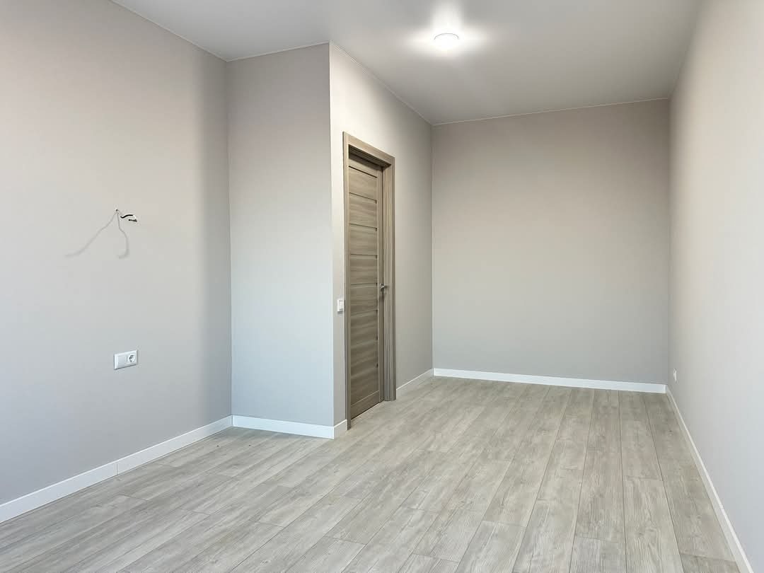 Pusty pokój po remoncie: świeżo pomalowane szare ściany, jasna drewniana podłoga, białe listwy przypodłogowe i drewniane drzwi. Widoczne gniazdko i wystające kable.