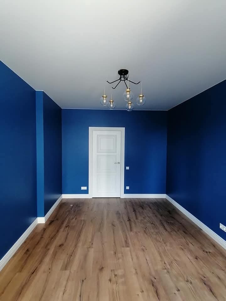 Pomalowane na intensywny niebieski pokój z drewnianą podłogą i nowoczesną lampą sufitową. Białe listwy przypodłogowe i drzwi tworzą kontrast.