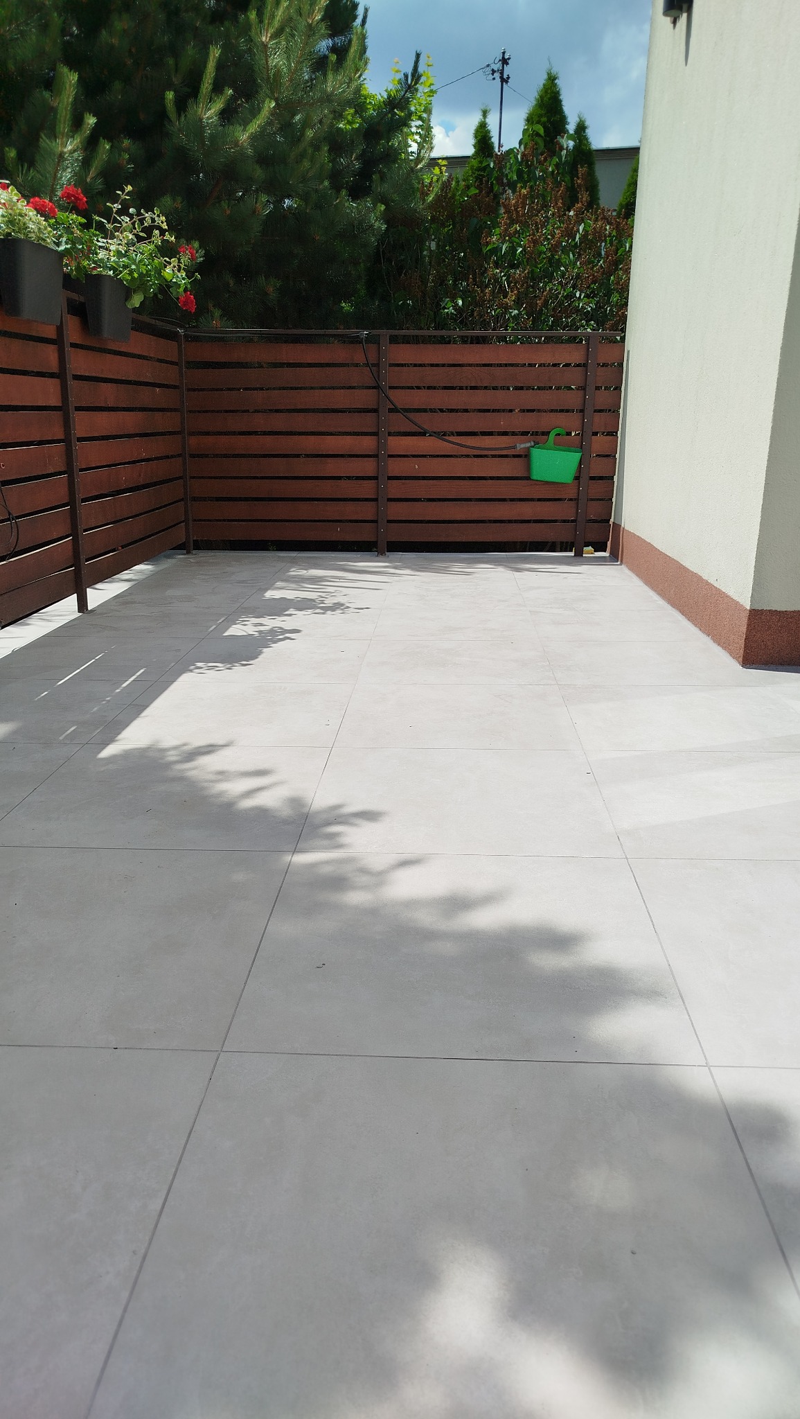 Jasne, kwadratowe płytki tarasowe na zewnątrz, otoczone drewnianym ogrodzeniem z roślinami i zielonym pojemnikiem, przylegające do ściany budynku z brązowym cokołem.