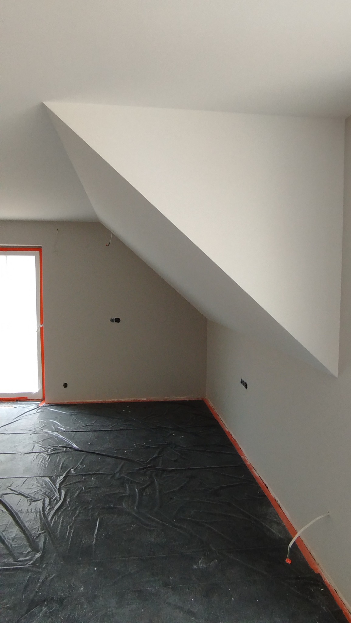 Poddasze po szpachlowaniu, przygotowane do malowania. Zabezpieczona folią podłoga, ściany w jasnym odcieniu, widoczne punkty elektryczne. Skos dachu.