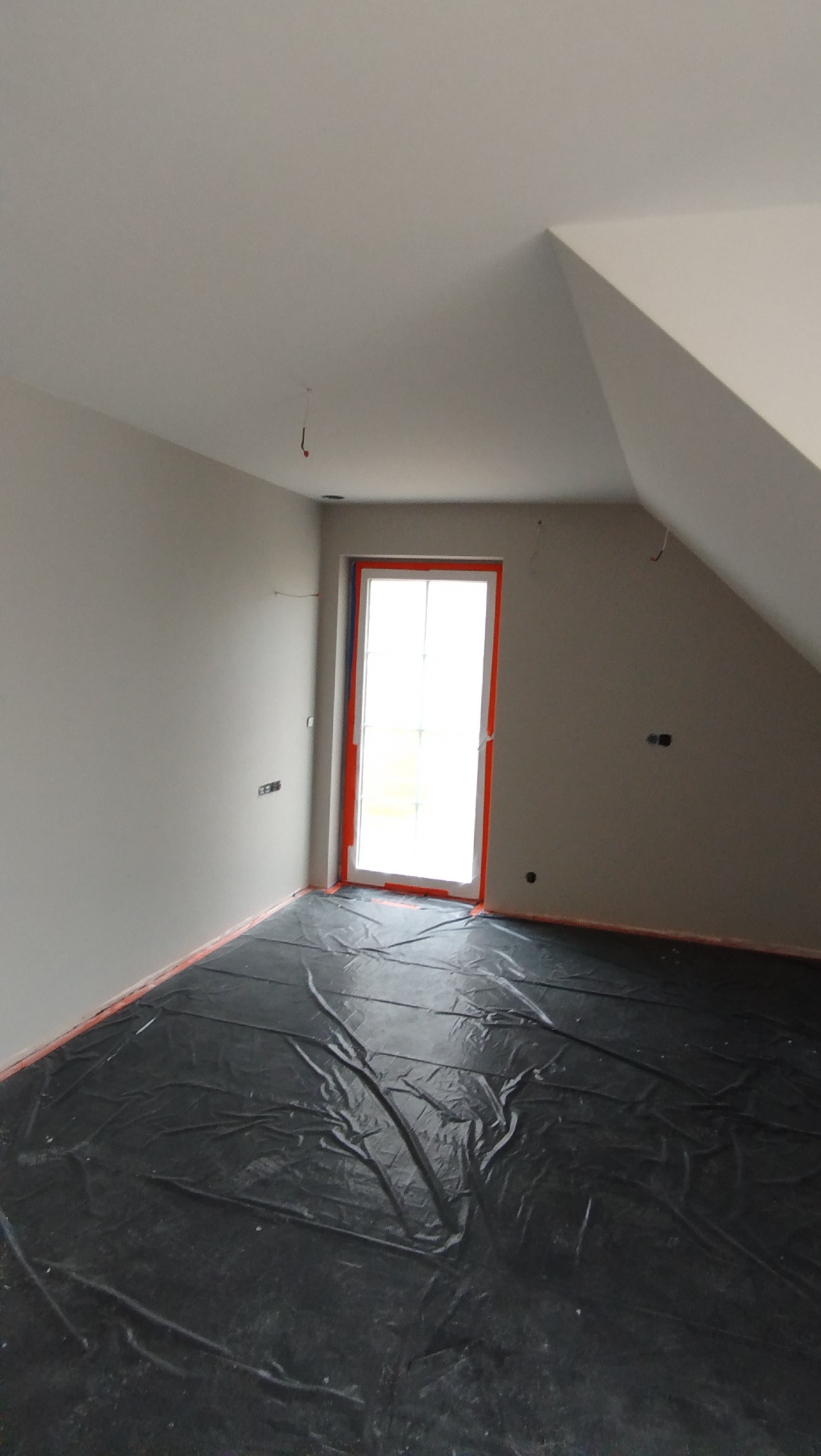 Przygotowane do prac wykończeniowych poddasze w Warszawie. Zabezpieczona folią podłoga, ściany po wstępnym malowaniu, okno z pomarańczową ramą.