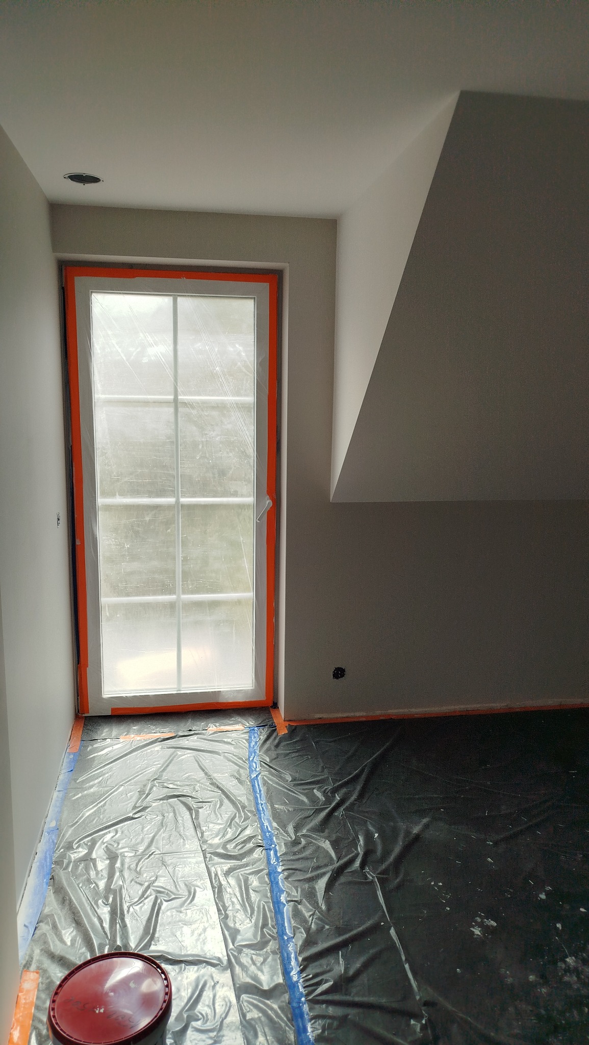 Pokój na poddaszu w trakcie przygotowań do malowania: okno zabezpieczone folią, podłoga przykryta czarną folią ochronną, ściany świeżo pomalowane na szaro.