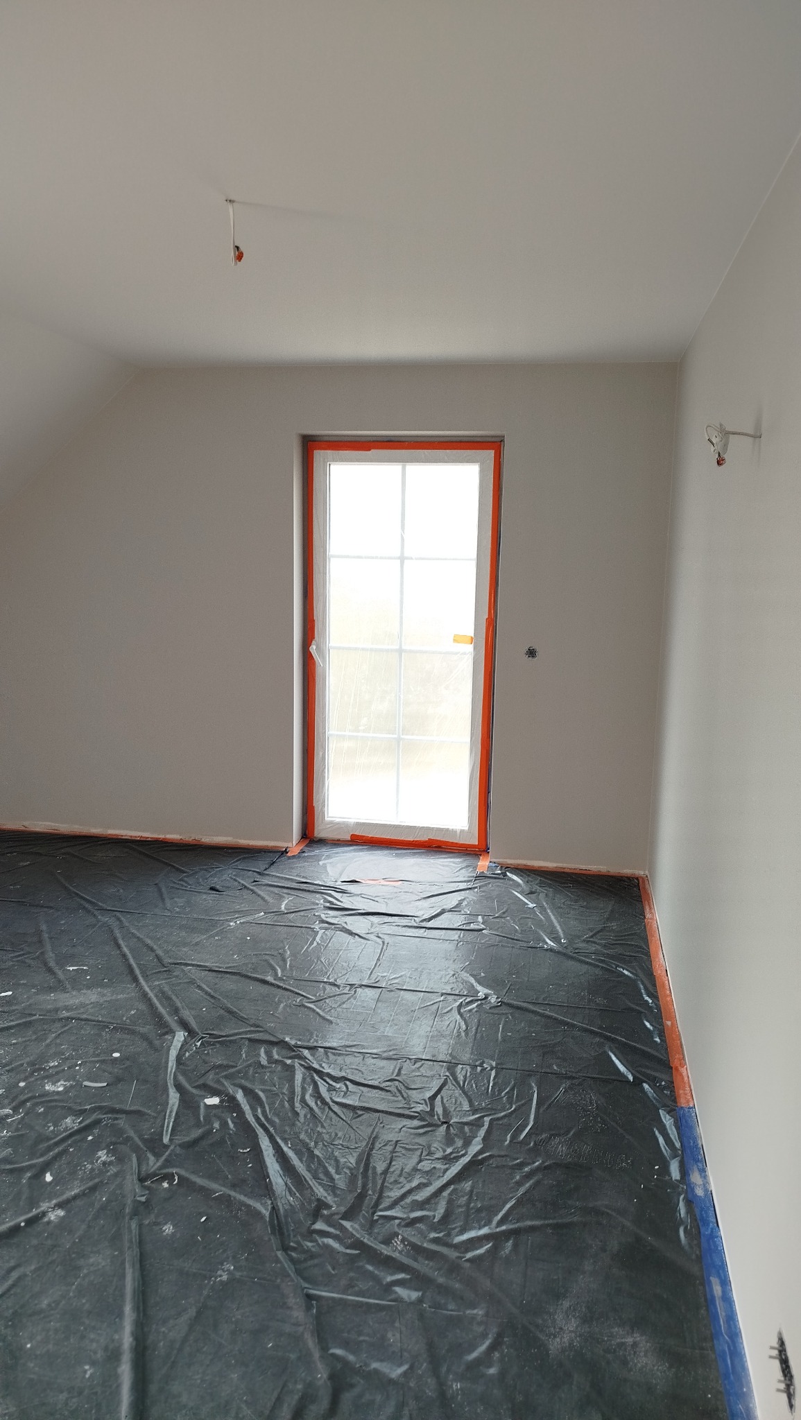 Pokój po szpachlowaniu, przygotowany do malowania. Okno zabezpieczone folią, podłoga przykryta czarną folią ochronną. Ściany gładkie, białe.