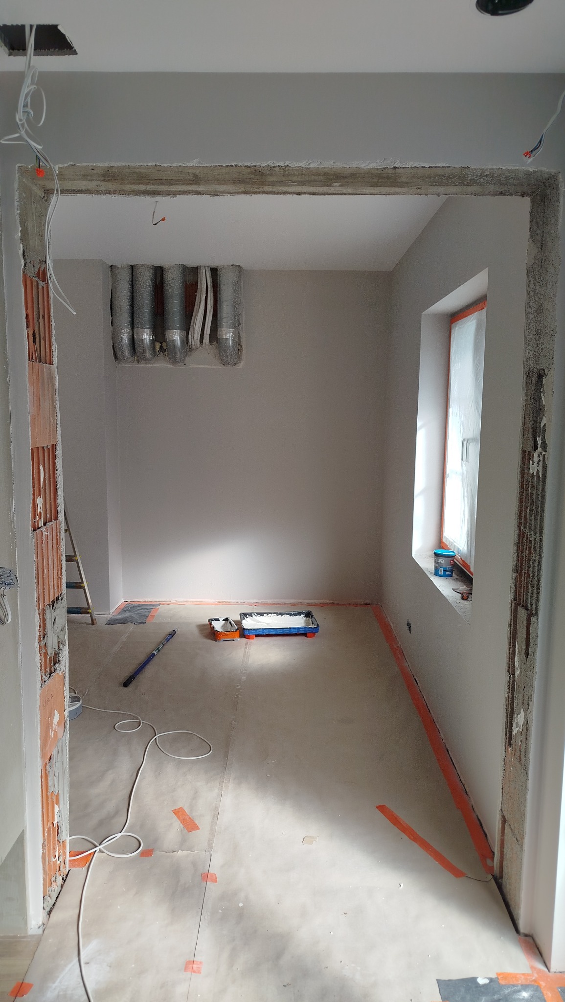 Pokój po malowaniu ścian na szaro, widoczne zabezpieczone okno, narzędzia malarskie i instalacja wentylacyjna. Przygotowanie do dalszych prac wykończeniowych.