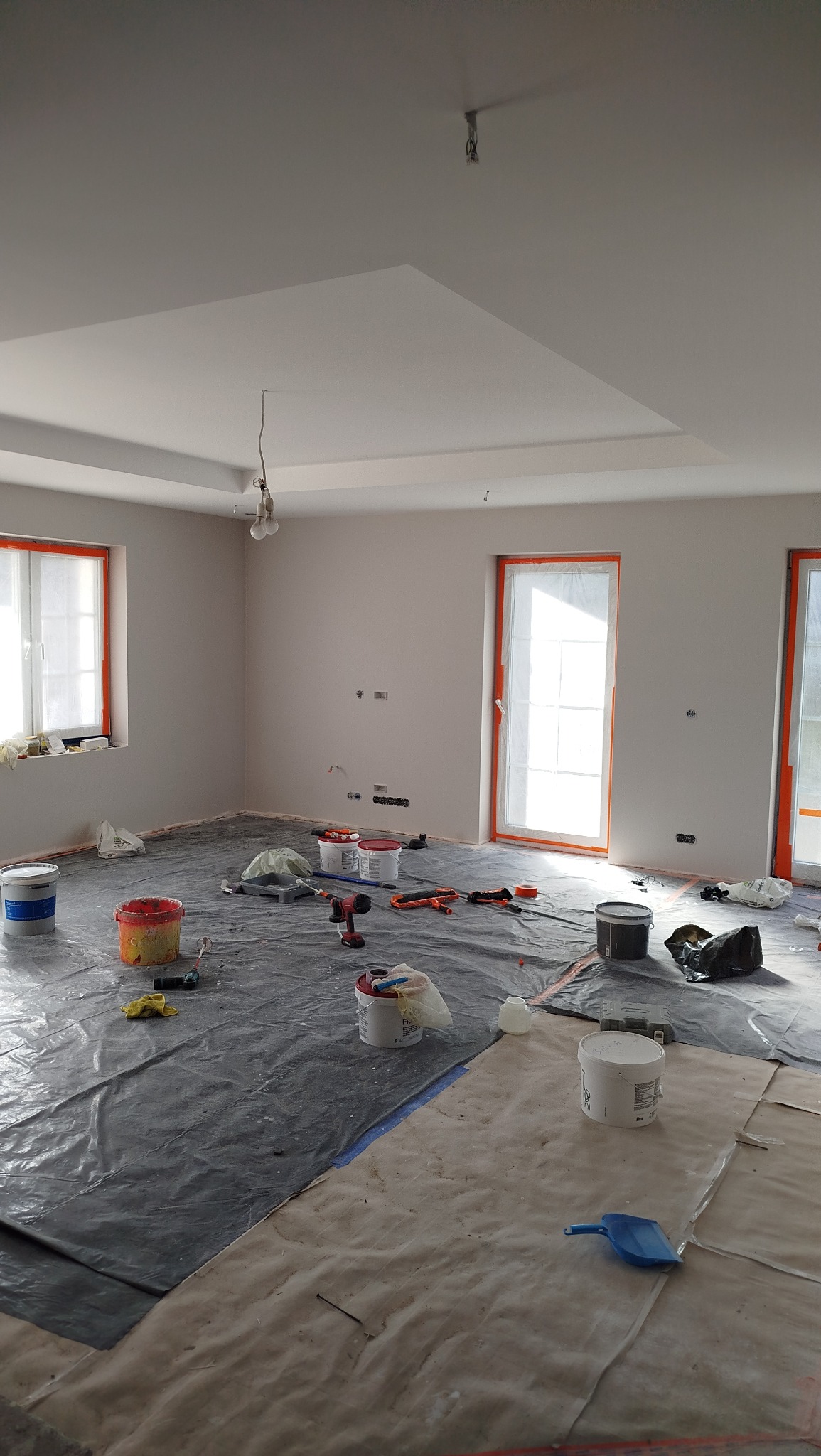 Pokój w trakcie malowania: zabezpieczona folią podłoga, pędzle, wałki, farby. Świeżo pomalowane ściany na jasny odcień. Okna i drzwi zabezpieczone folią malarską.