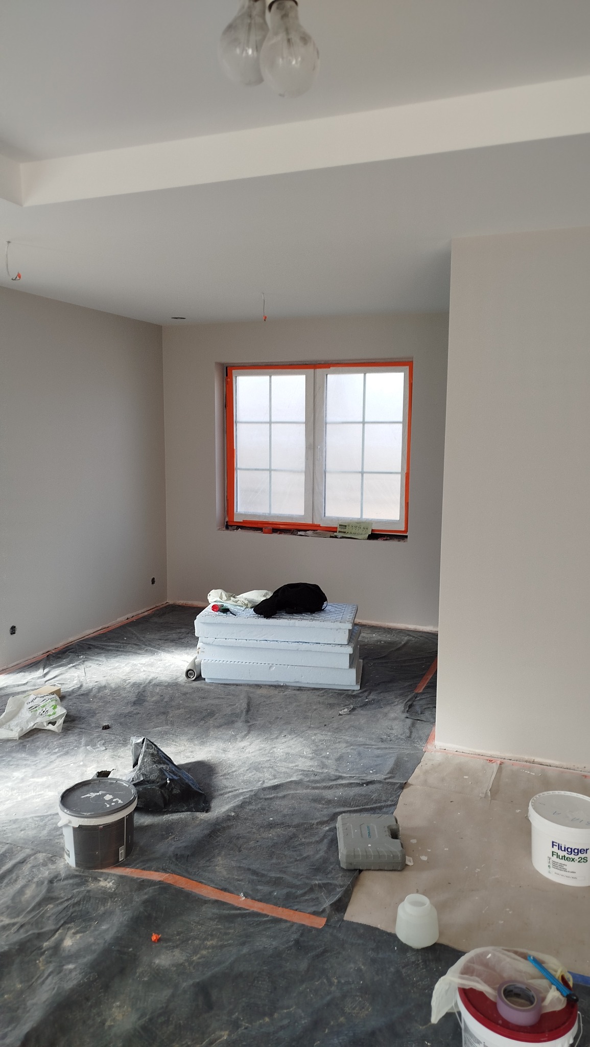 Wnętrze w trakcie prac wykończeniowych: zabezpieczona folią podłoga, puszki z farbą, styropian przy oknie. Świeżo pomalowane ściany w jasnym odcieniu.