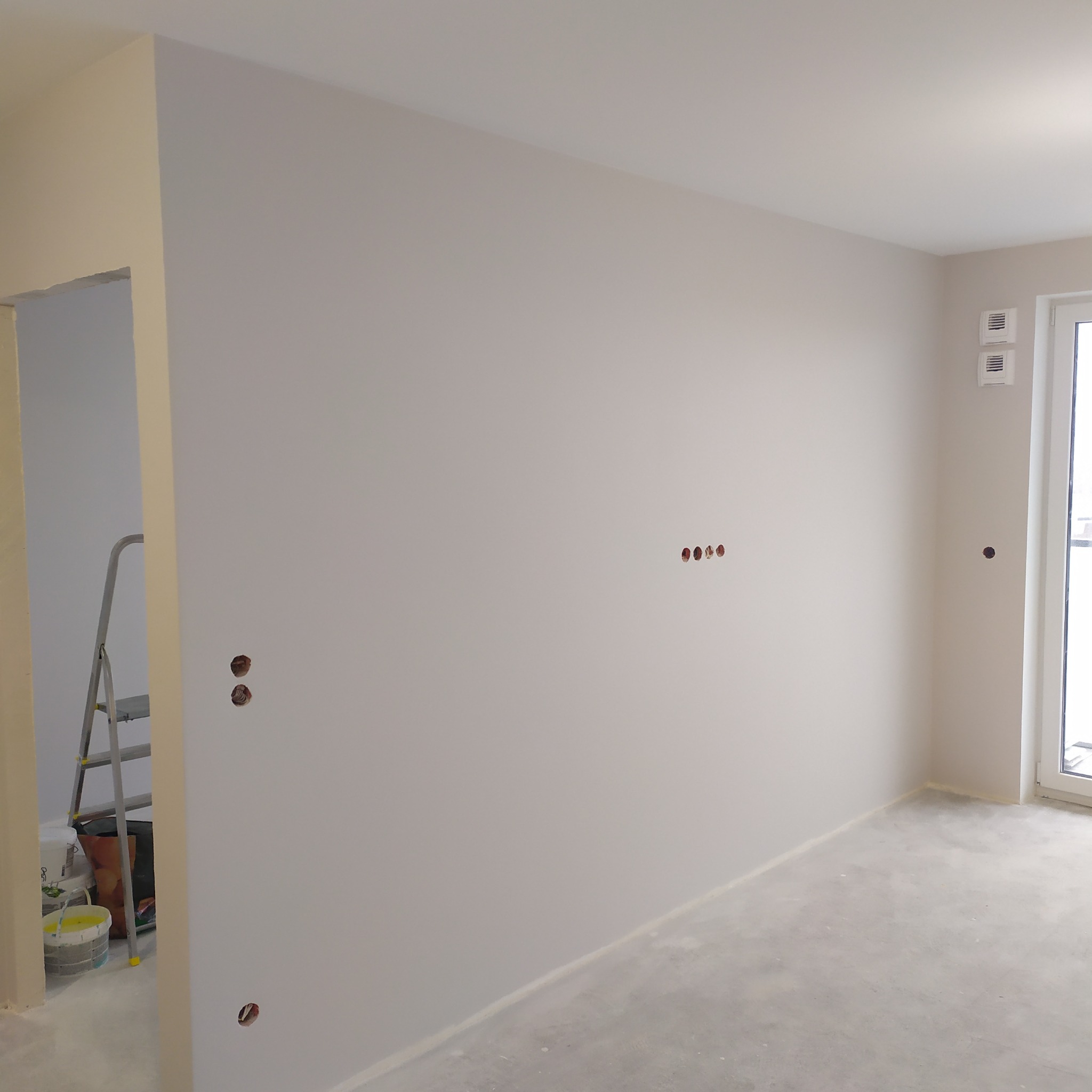 Jasne, świeżo pomalowane wnętrze z widocznymi puszkami elektrycznymi i drabiną w tle, sugerujące etap prac wykończeniowych. Minimalistyczny styl.