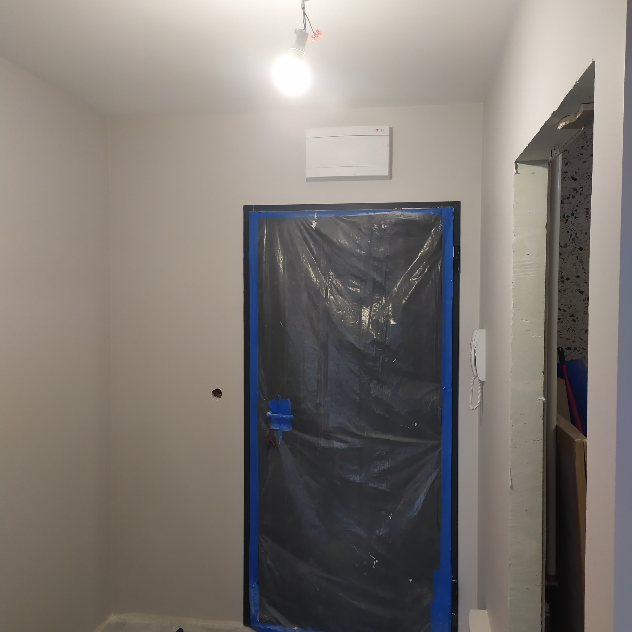 Przygotowanie do malowania: zabezpieczone folią drzwi z niebieską taśmą malarską, świeżo pomalowana ściana w odcieniach szarości, widoczna instalacja elektryczna.