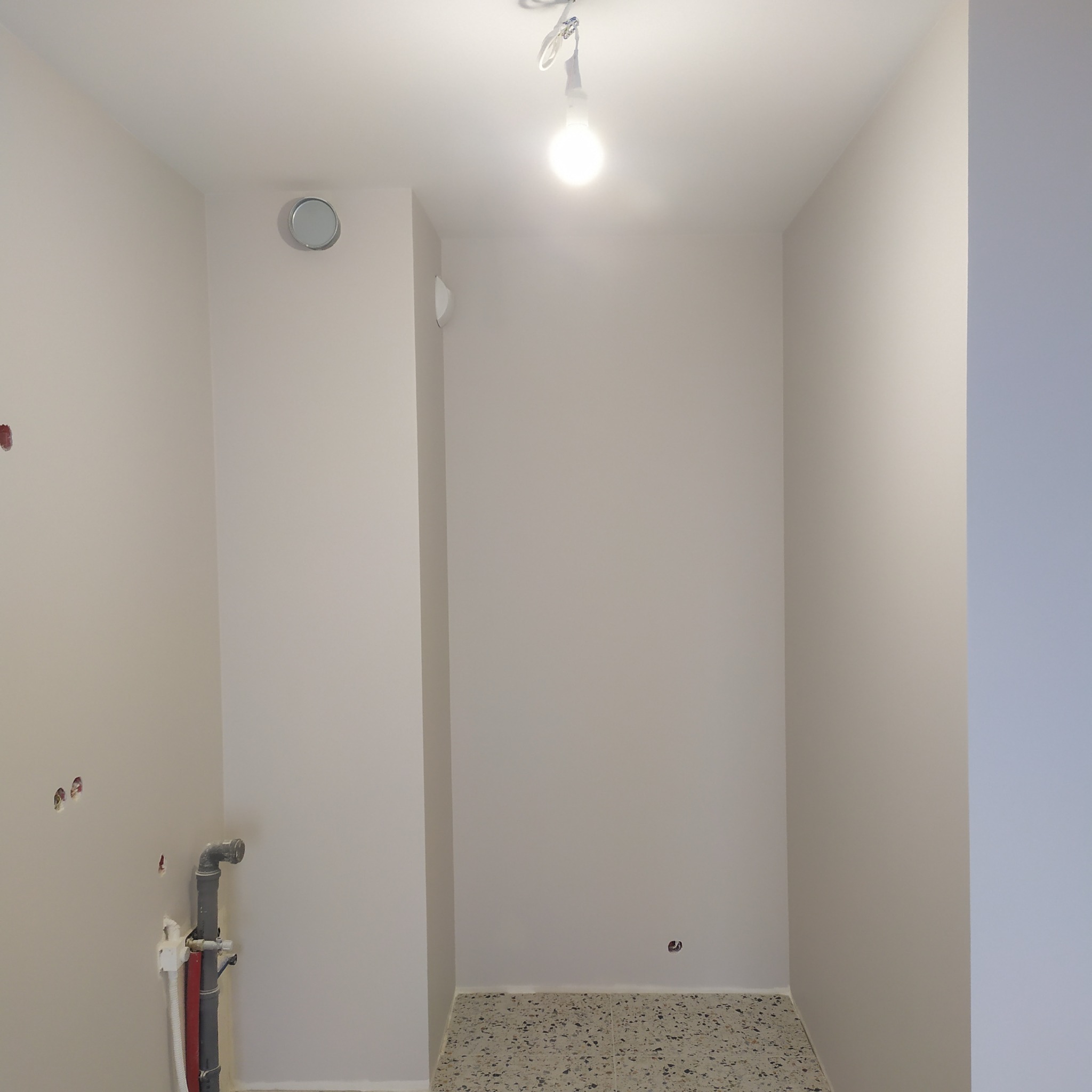 Wnętrze po odświeżeniu: gładkie, jasne ściany w odcieniach bieli, widoczne rury instalacyjne i fragment posadzki z lastryko. Minimalistyczne oświetlenie sufitowe.